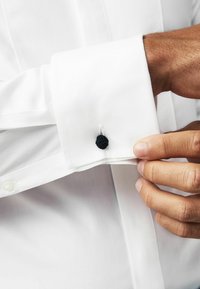 Weißes Hemd aus strukturiertem Stoff, ausgestattet mit einer schwarzen Krawattenmanschette. Nahaufnahme des Manschettendetails und einer Hand, die die Manschette anpasst.