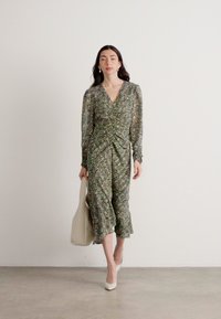 Robe portefeuille à fleurs dans des nuances de vert, dotée de manches longues, d'une taille cintrée et d'une silhouette fluide. Associée à un sac à main crème et des talons.