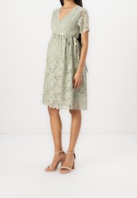 Vihreä pitsimekko, jossa on V-pääntie, lyhyet hihat ja sidottu vyötärö. Värivarvelinen helma lisää tekstuuria. Päällä luonnonväriset korkokengät.