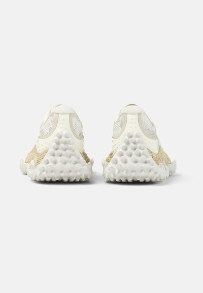 Pár béžových a bílých tenisek s texturovanými, zaoblenými podrážkami zobrazených zezadu na jednoduchém světlém pozadí.