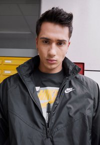 Melna neilona jaka ar augstu apkakli, ar rāvējslēdzēju priekšā un Nike logo. Zem jakas ir pelēks T-krekls ar dzeltenu grafisku dizainu. Īsi ieveidoti mati.
