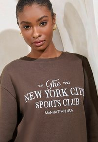 Felpa in cotone marrone con scritta bianca: "The New York City Sports Club Manhattan USA" fondata nel 1993; vestibilità comoda, scollo rotondo.
