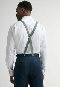 Tirantes verdes con clips plateados sobre una camisa blanca a rayas, combinados con pantalones azul marino con bolsillos traseros y un detalle de botón.