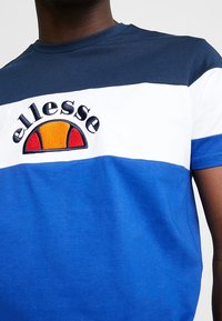 Muž v modrém, bílém a námořnickém barveném tričku s bloky barev a vyšitým oranžovým a červeným logem ellesse na hrudi.