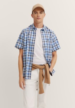 Kurzärmliges, kariertes Hemd in Blau, Weiß und Braun mit einer knöpfbaren Front. Darunter ein weißes T-Shirt und ein brauner Pullover, der um die Taille gebunden ist.