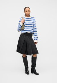 Γυναίκα που φοράει πουλόβερ με μπλε και λευκές ρίγες, μαύρη πλισέ φούστα, μαύρες μπότες μέχρι το γόνατο και κρατά μια μικρή τσάντα ελιάς πράσινου χρώματος.