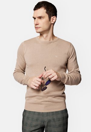 Beżowy sweter dzianinowy z okrągłym dekoltem i prążkowanymi mankietami, trzymany w ręce z okularami przeciwsłonecznymi. Sweter posiada małe logo na piersi.