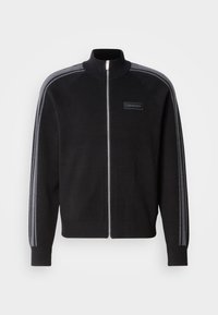 Zwarte jas met ritssluiting en hoge kraag, grijze gestreepte accenten op de mouwen en een label aan de voorkant. Gemaakt van een gladde stoftextuur.