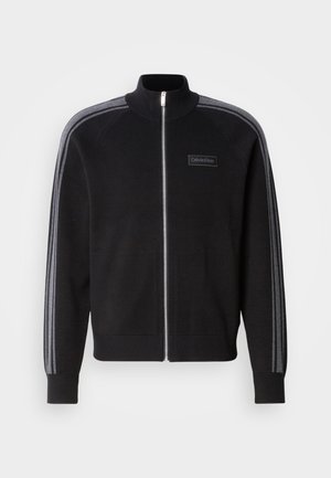 Casaco preto com fecho de correr e gola alta, detalhes em riscas cinzentas nas mangas e etiqueta frontal. Feito de um tecido com textura suave.