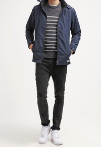 La giacca navy presenta una chiusura con zip, colletto alto e cucitura sul petto. Sotto, si intravede un maglione grigio a righe. Abbinato a pantaloni neri e sneaker bianche.
