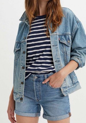 Kvinde iført blå denimjakke og shorts med navy og hvidstribet skjorte, hånd i lommen, mod ensfarvet baggrund.