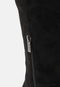Stivale in suede nero al ginocchio con zip laterale e un tirante in argento. La texture è liscia con una leggera lucentezza.