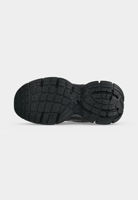 Semelle de chaussure en caoutchouc noir avec des motifs de bandes de roulement rectangulaires et courbes texturés pour l’adhérence, montrée sur un fond clair uni.