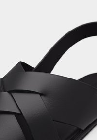 Μαύρο δερμάτινο σανδάλι με φαρδιές λουρίδες που διασταυρώνονται και λουρί στο πίσω μέρος, απεικονίζεται σε κοντινή λήψη σε απλό ανοιχτό φόντο.