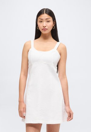 Kvinde med langt mørkt hår iført guld hoop øreringe og en ærmeløs hvid kjole, der står mod en ensfarvet lys baggrund.