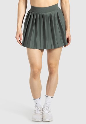 Falda plegada verde hecha de tela suave con cintura alta. La longitud es por encima de la rodilla, combinada con calcetines y zapatos deportivos blancos.