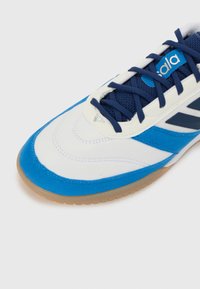 Zapatilla deportiva blanca y azul con parte superior de ante y malla, que presenta una suela de goma y acentos azules en los lados y los cordones.