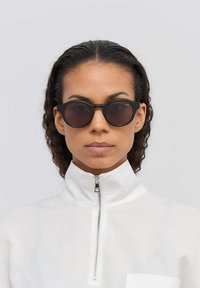 Pyöreät mustat aurinkolasit tummilla linsseillä, mattapintaiset ja minimalistisella muotoilulla. Mallilla on päällään valkoinen vetoketjullinen takki ja kiharat hiukset.