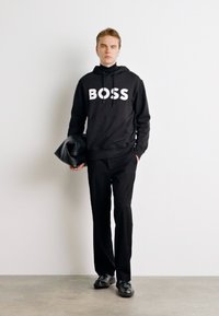 Juodas džemperis su baltu "BOSS" logotipu, derinamas su juodomis kelnėmis ir bateliais. Modelis laiko juodą rankinę, stovėdamas prieš neutralią fono.