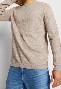Man som bär en beige tröja med rund halsringning, med en liten röd, vit och blå logotyp på bröstet, samt blå jeans, poserande med en arm böjd nära ansiktet.