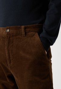 Brune corduroy bukser med en struktureret overflade, der har knaplukning, bæltestropper og lommer. Sammen med en mørk sweater.