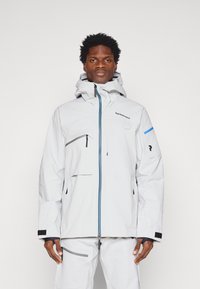 Chaqueta blanca impermeable con capucha, detalles en azul, bolsillos con cremallera, puños ajustables y un logo en el pecho. Material liso y funcional.