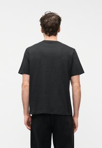 Άνδρας με κοντά καστανά μαλλιά, γυρισμένος πλάτη, φο ράει απλό μαύρο κοντομάνικο μπλουζάκι και μαύρα τζιν, σε λευκό φόντο.