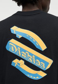 Άτομο που φοράει μαύρο πουκάμισο με μεγάλο τρισδιάστατο κίτρινο και μπλε λογότυπο "Dickies" στην πλάτη.