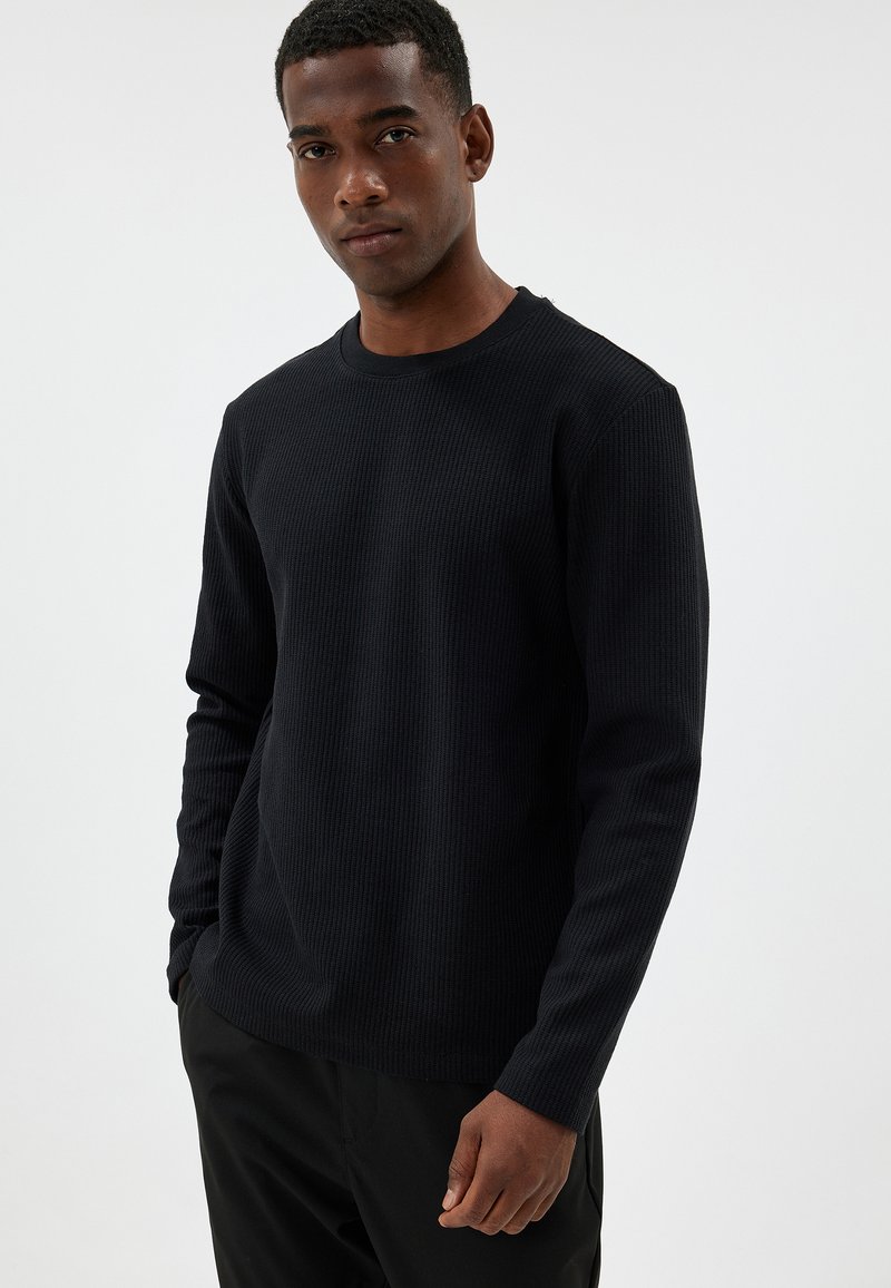 Schwarzes, geripptes Langarmshirt mit Rundhalsausschnitt, aus strukturiertem Stoff und mit enganliegenden Bündchen, kombiniert mit dunklen Hosen.