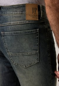 Vaqueros de mezclilla oscura con un acabado desgastado, presentando una etiqueta de cuero marrón en la cinturilla y dos bolsillos traseros con costuras sutiles.