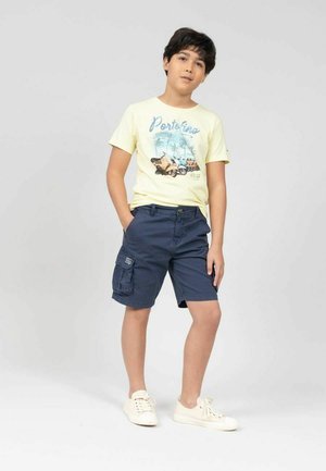 T-shirt jaune clair avec des impressions de paysages, associé à un short cargo bleu marine. L'ensemble est complété par des baskets blanches.