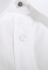 Tessuto di camicia bianca con colletto appuntito. Presenta un bottone bianco rotondo e un accento decorativo argentato sopra il bottone. Texture liscia.