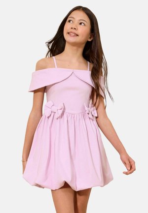 Girl in a light pink off-shoulder dress with thin straps, two bow accents at the waist, and a voluminous skirt, looking upward.