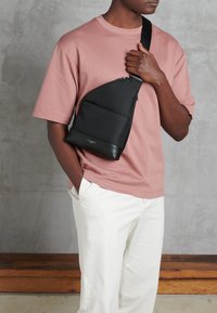 Borsa a tracolla nera con una forma affusolata, realizzata in pelle texturizzata, dotata di una tasca frontale con zip e tracolla regolabile, indossata su una maglietta rosa.