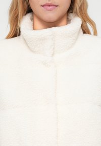 Bela flis jakna z visokim ovratnikom, prešitjem in mehkobo, s vidnimi šivi in minimalističnim estetskim izgledom.