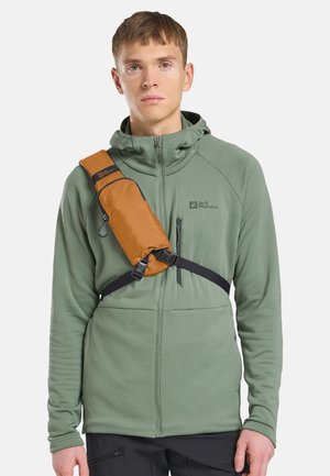 Junger Mann trägt eine hellgrüne Reißverschlussjacke und schwarze Hose, mit einer kleinen braunen Umhängetasche mit schwarzem Riemen quer über der Brust.