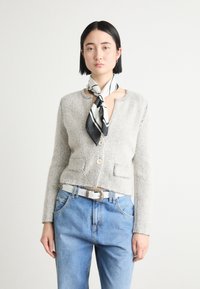 Lichtgrijze visgraat cardigan met lange mouwen, twee zakken en een ronde halslijn, gedragen over blauwe jeans en geaccentueerd met een zwart-wit sjaal.