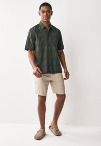 Dunkelgrünes T-Shirt mit kurzem Arm und strukturiertem Muster, kombiniert mit beigen Leinen-Shorts und grauen Slip-on-Schuhen. Modell steht vor neutralem Hintergrund.