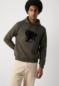 Haut en coton vert olive avec cordon de serrage, présentant un grand motif graphique noir à l'avant et un ourlet côtelé. Le modèle porte un pantalon beige.