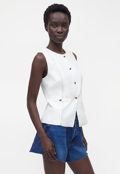 Femme portant un gilet blanc sans manches boutonné avec des boutons dorés et un short en jean bleu, debout devant un fond blanc uni.