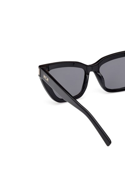 Černé sluneční brýle ve stylu cat-eye se šedými skly, lesklým povrchem a zlatým logem "MCM" na spánku. Disponují moderním úhlovým designem.