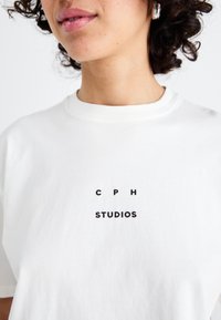 Hvid bomulds t-shirt med teksten "CPH" i sorte store bogstaver og "STUDIOS" nedenunder i fed skrift. Rund halsudskæring.