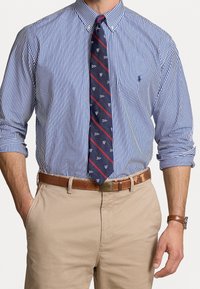 Man som bär en blå och vit randig skjorta med uppvikta ärmar, marinblå mönstrad slips, beige byxor samt brunt läderbälte och klocka.