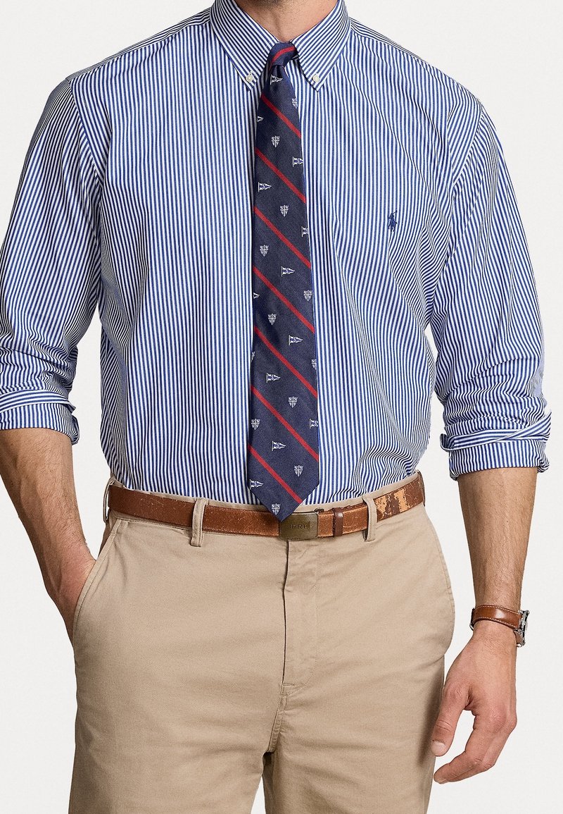 Man som bär en blå och vit randig skjorta med uppvikta ärmar, marinblå mönstrad slips, beige byxor samt brunt läderbälte och klocka.