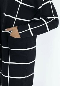 Cardigan noir avec un motif à carreaux blancs, fabriqué dans un matériau doux. Caractéristiques : manches longues et poches latérales.