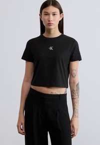 Μαύρο cropped t-shirt από βαμβάκι, με λευκό κεντημένο λογότυπο στο στήθος, συνδυασμένο με μαύρο παντελόνι tailored. Λεία υφή.