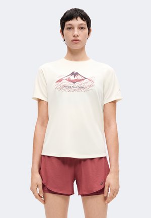 Personne portant un t-shirt blanc avec un motif de montagne et un short rouge, debout face à un fond uni.