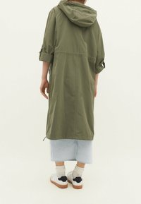 Olive green, long, hooded jacket with rolled sleeves, made of lightweight material. Worn over a light denim skirt and white sneakers.