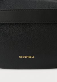 Sac en cuir noir avec une surface texturée et une bande lisse. "COCCINELLE" embossé en or en bas. Design minimaliste.