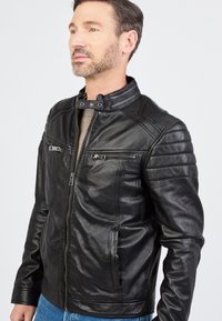 Schwarze Lederjacke mit hohem Kragen, Reißverschluss vorne und mehreren Reißverschluss-Taschen. Glatte Textur mit gesteppten Schulterakzenten.